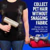 A person holding the roller against a black t-shirt covered in shed fur to show it cleans without snagging fabric. A small dog rests in a red suitcase in the corner